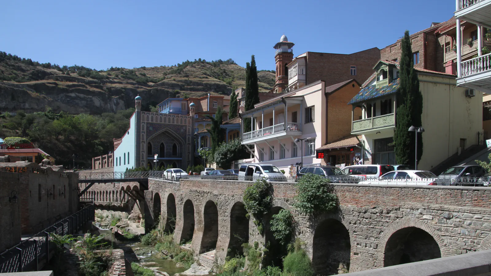 Kamnit in opečnat obokan zid nad potokom v četrti Abanotubani, za hišami modro obloženo pročelje kopališča in opečni minaret