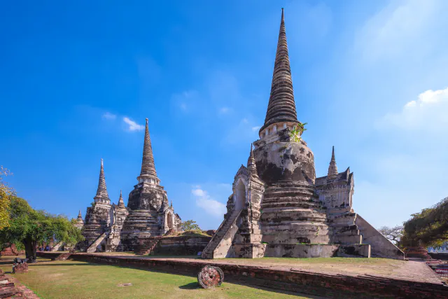 Tri stupe z visokimi koničastimi vrhovi in obrabljenimi sivimi ter rjavimi površinami v templju Wat Phra Si Sanphet v Ayutthayi pod modrim nebom