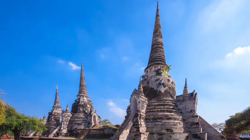 Tri stupe z visokimi koničastimi vrhovi in obrabljenimi sivimi ter rjavimi površinami v templju Wat Phra Si Sanphet v Ayutthayi pod modrim nebom