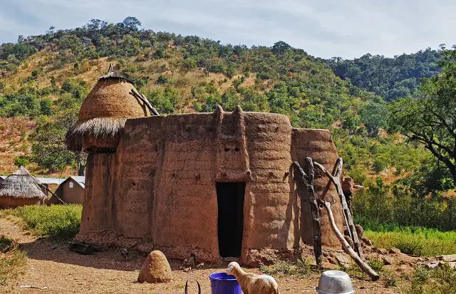 Batammariba tower-house with a thatched turret, set against a green wooded hillside.