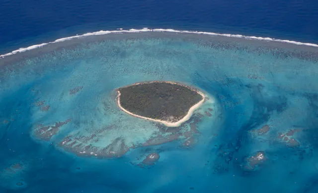 Aerial view of a small wooded island with a pale sandy shoreline, surrounded by shallow turquoise reef water; surf breaks along the outer reef beyond it.
