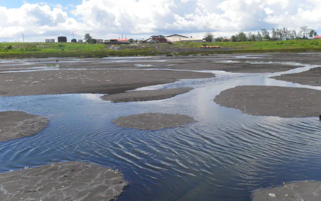 Temne zaplate naravnega asfalta med plitvimi bazenčki z nagubano vodno gladino na jezeru Pitch Lake pri La Brei na Trinidadu; v ozadju so nizke stavbe, zelen pas trave in oblačno nebo.