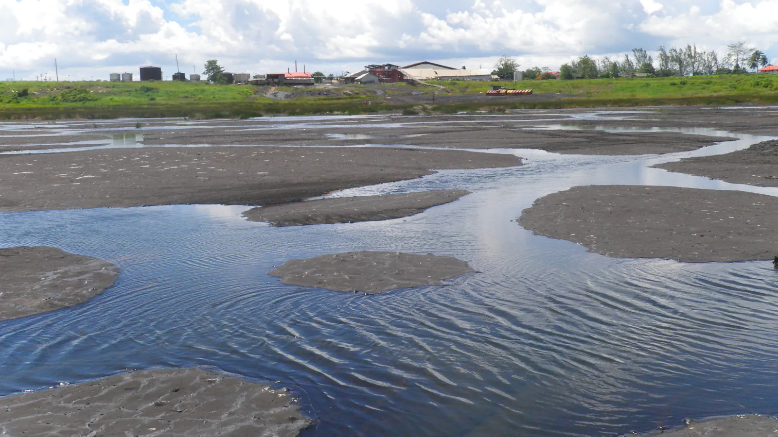 Temne zaplate naravnega asfalta med plitvimi bazenčki z nagubano vodno gladino na jezeru Pitch Lake pri La Brei na Trinidadu; v ozadju so nizke stavbe, zelen pas trave in oblačno nebo.