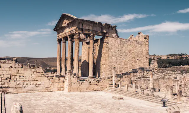 Columned front of the Capitol temple at Dougga rising over ruined walls, with a paved square in front and dry hills behind under a blue sky