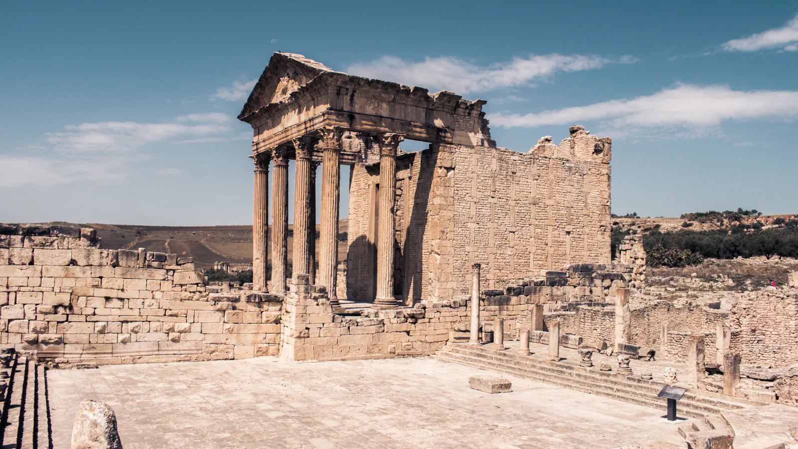 Columned front of the Capitol temple at Dougga rising over ruined walls, with a paved square in front and dry hills behind under a blue sky