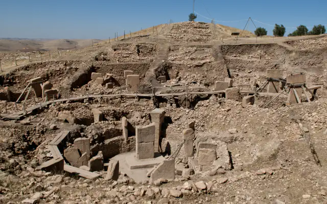Excavated stone structures with T-shaped pillars on a dry, bare-looking Göbekli Tepe hillside under a clear sky