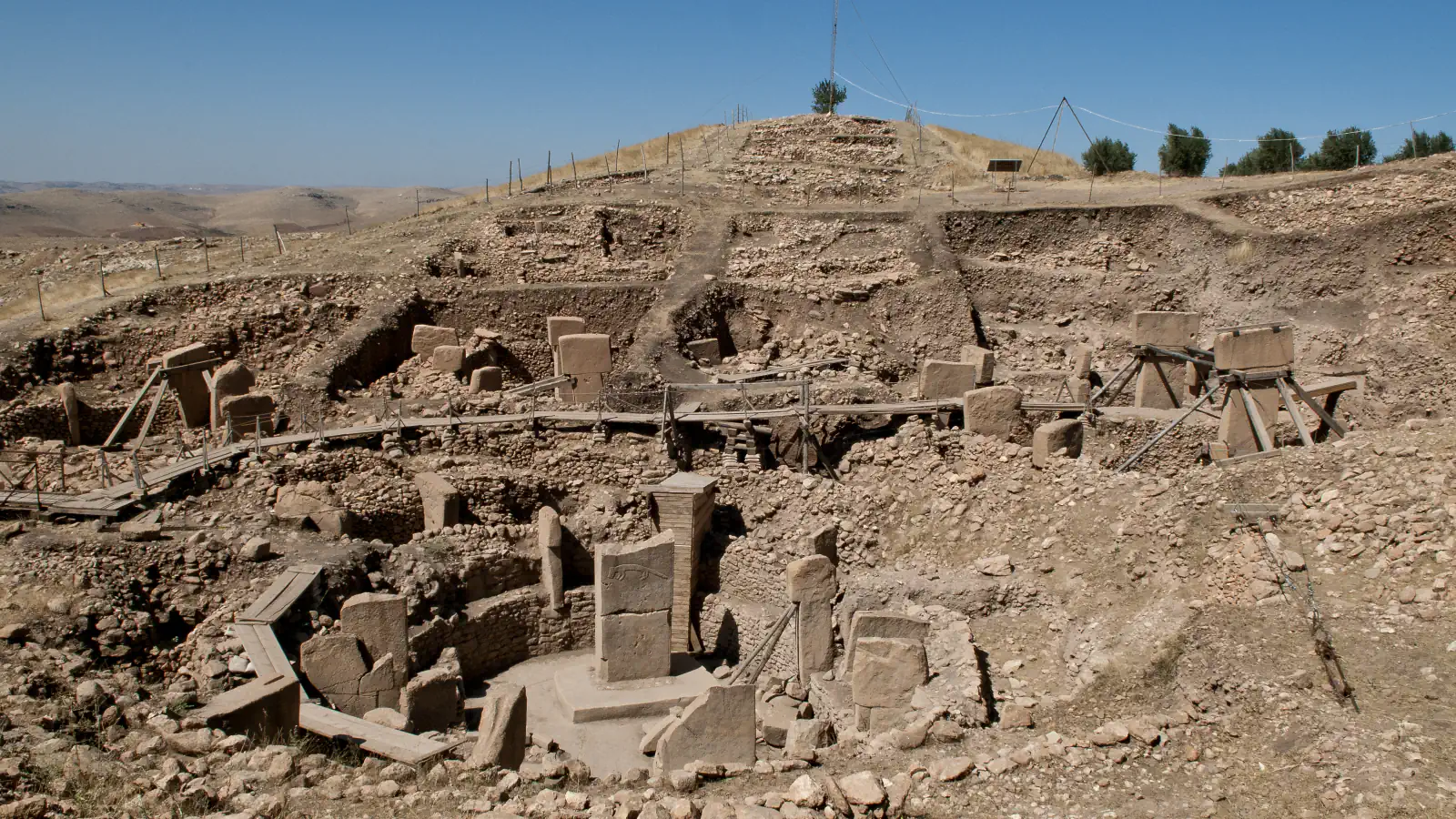 Excavated stone structures with T-shaped pillars on a dry, bare-looking Göbekli Tepe hillside under a clear sky