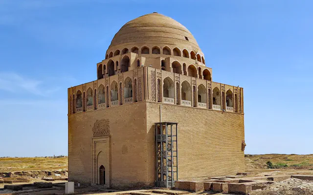 Square brick mausoleum with arched galleries below a ribbed dome and a steel stair tower on one side, standing on desert ground among low excavated walls