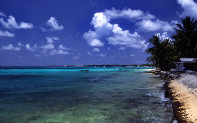 Shoreline at Funafuti with turquoise water, a small boat, coconut palms and low buildings; a sloping concrete seawall runs along the right beneath a clouded blue sky.