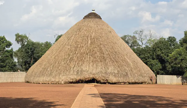 A large conical building thatched with grass and topped by a small pointed finial, seen from a courtyard of red earth with a brick path leading to its low entrance, with trees and a fence of vertical stalks behind it