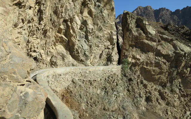 A narrow water channel with concrete edges curves along a steep, rocky mountainside, with a strip of blue sky above the cliffs