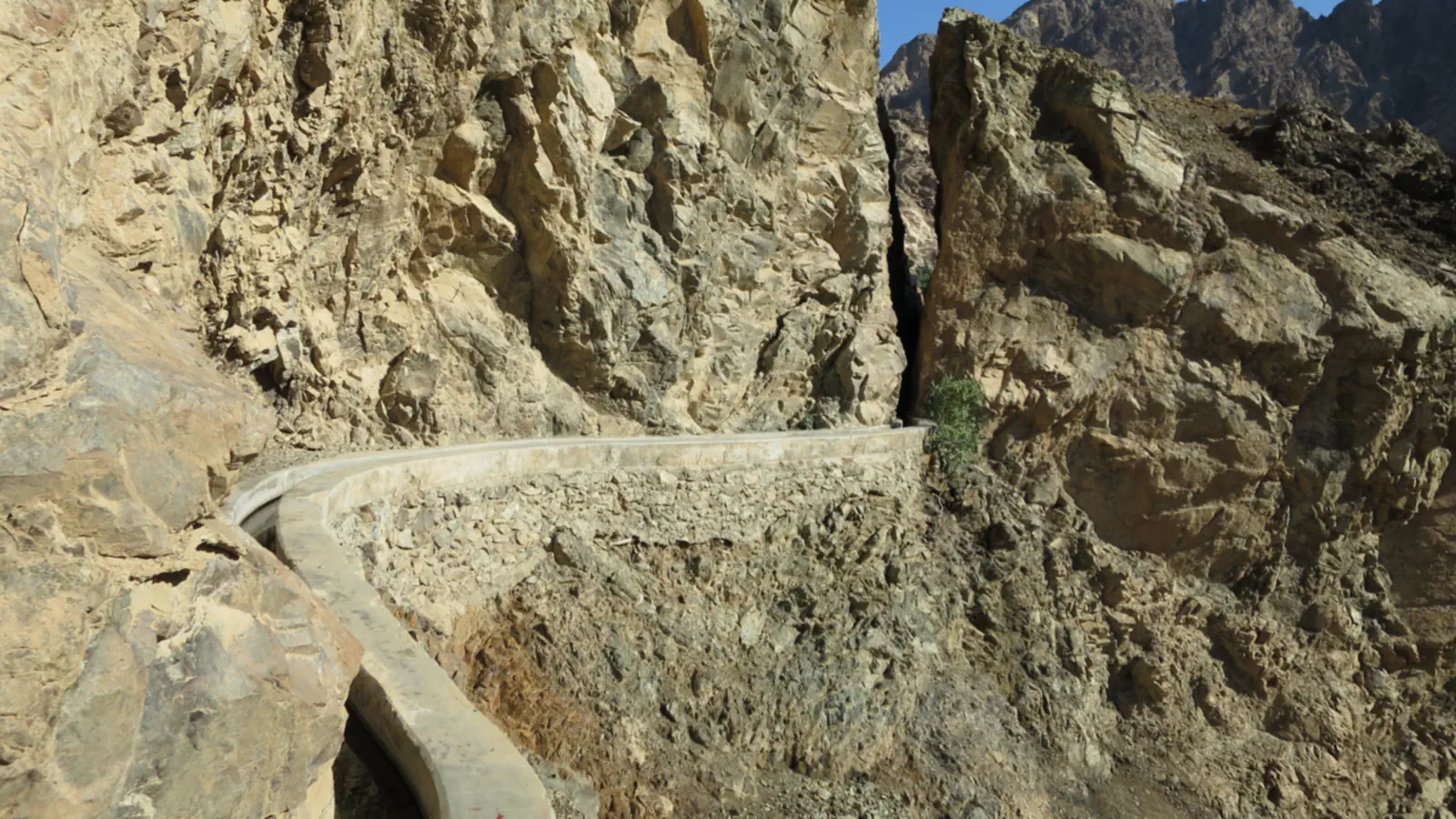 A narrow water channel with concrete edges curves along a steep, rocky mountainside, with a strip of blue sky above the cliffs