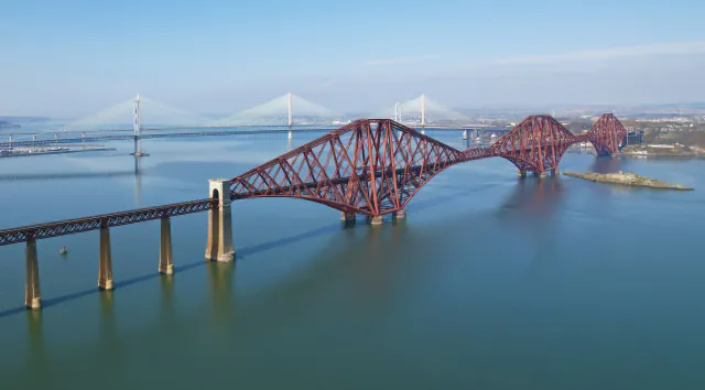 Aerial view of the red railway span over the Firth of Forth, with road bridges visible beyond.