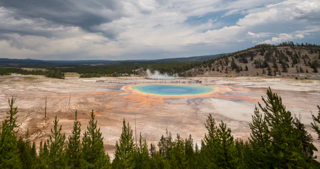 Grand Prismatic Spring, okrogel turkizen bazen z oranžnim in rumenim robom, viden s pobočja pod nevihtnim nebom; spredaj borovci, za bazenom lesena pot