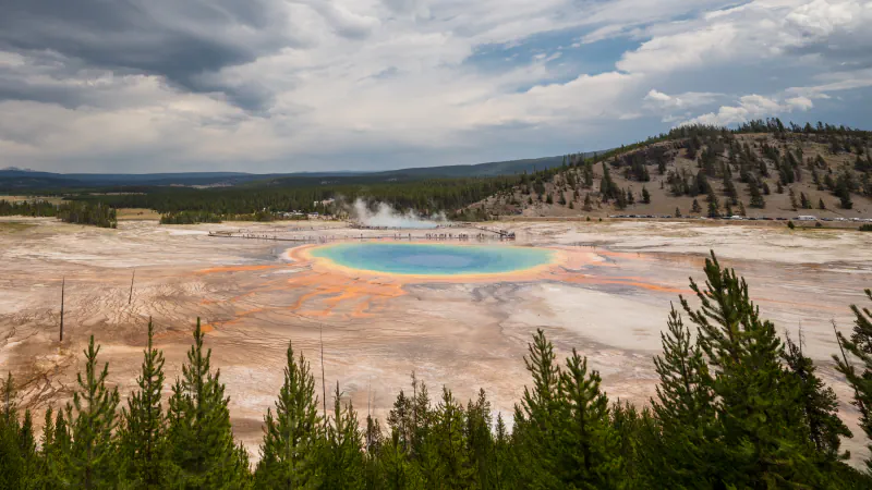 Grand Prismatic Spring, okrogel turkizen bazen z oranžnim in rumenim robom, viden s pobočja pod nevihtnim nebom; spredaj borovci, za bazenom lesena pot