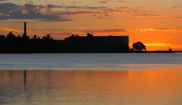 Silhouette of the former Anglo meat plant at sunset on the Uruguay River: a tall chimney at the left, a large dark industrial building in the centre and a crane at the end of a pier on the right, all above orange-lit water