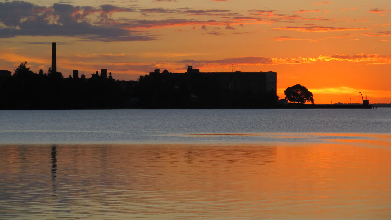 Silhouette of the former Anglo meat plant at sunset on the Uruguay River: a tall chimney at the left, a large dark industrial building in the centre and a crane at the end of a pier on the right, all above orange-lit water