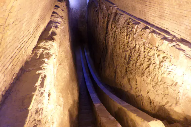 Looking down a narrow trench cut into pale earth, with two curved parallel stone-faced walls running along its floor and brick walls rising on either side