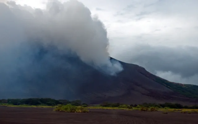 A dark, steep-sided volcanic cone rising behind a flat grey-black ash plain, with a thick plume of gas and cloud drifting across its upper slope and low green vegetation along the base