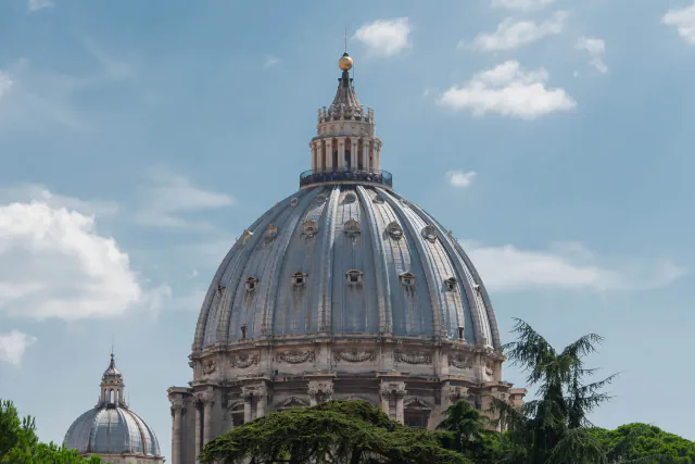 The grey-blue ribbed dome of St Peter’s Basilica with its lantern and golden ball, rising above dark green trees under a partly cloudy sky; a smaller dome stands at the lower left