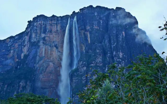 A tall, dark rock wall of a table mountain with two thin streams of water falling down its face, green forest in the foreground and low cloud under a pale blue sky