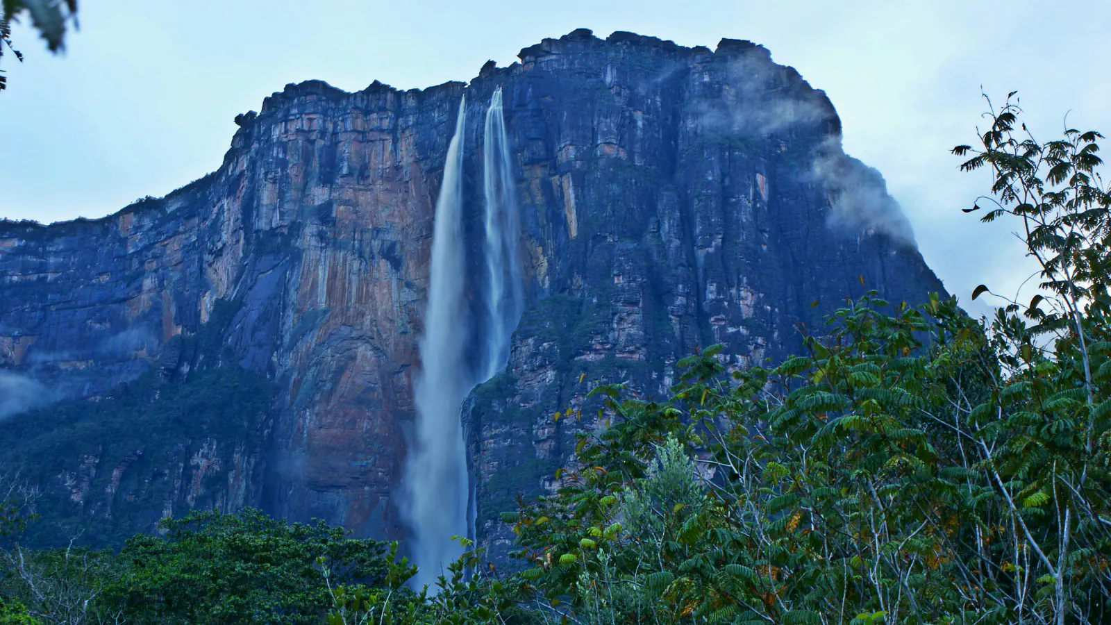 A tall, dark rock wall of a table mountain with two thin streams of water falling down its face, green forest in the foreground and low cloud under a pale blue sky