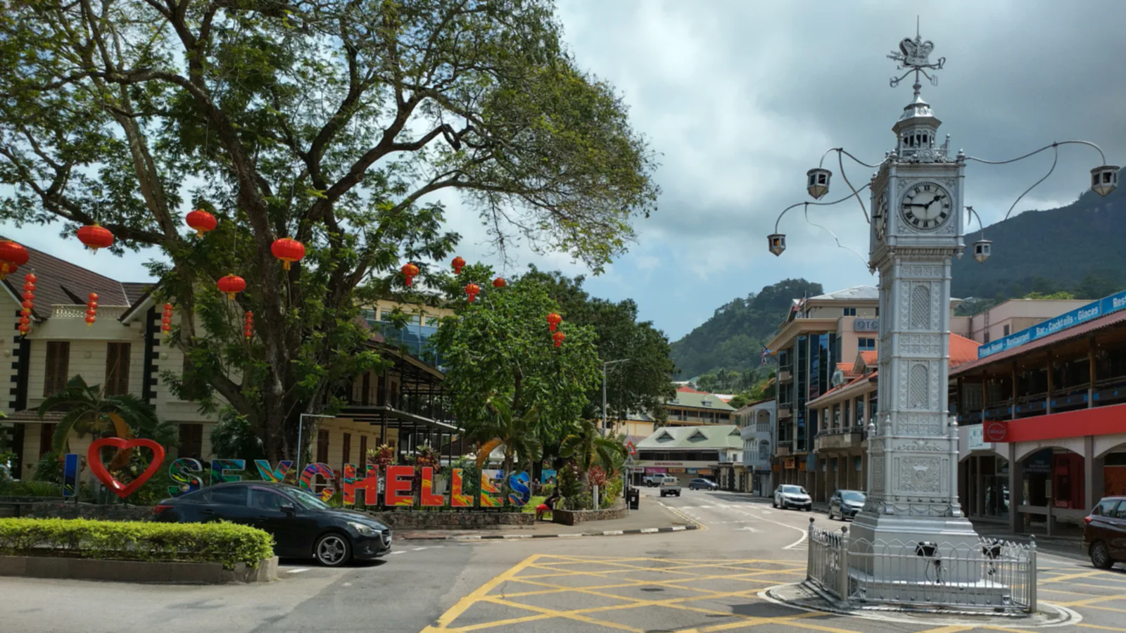 A silver-grey clock tower on a street corner, with a tree hung with red lanterns, a colourful lettered sign and green hills behind under a cloudy sky