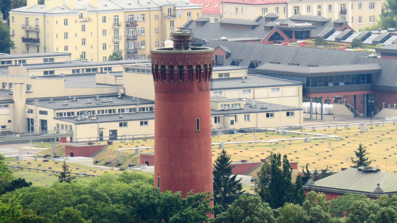 A red-brick water tower rising above the grass-covered roofs of filters, with low industrial buildings and blocks of flats behind.