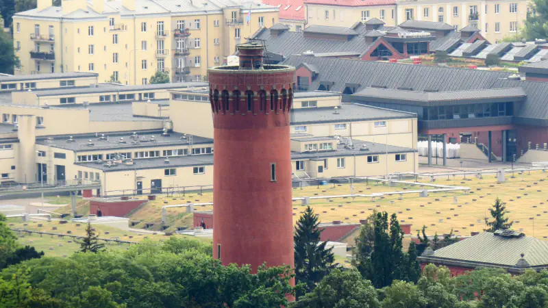 Rdeč opečnat vodni stolp nad zatravljenimi strehami filtrov, za njim nizke industrijske stavbe in stanovanjski bloki.