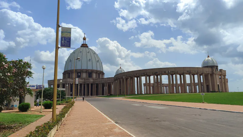 A large grey dome beside a curved colonnade ending in a smaller dome, seen from the road beneath a cloudy sky.