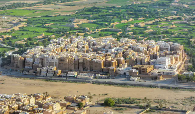 Aerial view of a dense cluster of tall mud-brick tower houses, with green irrigated fields beyond them and a wide, sandy wadi bed in the foreground