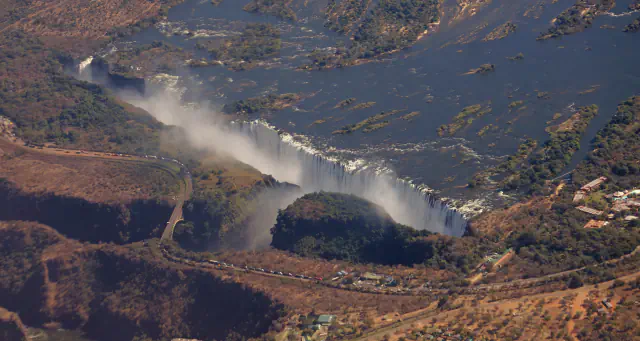 Aerial view of Victoria Falls: a long curtain of white water and mist along the lip of a deep gorge, with the Zambezi and its islands above and a road bridge crossing the gorge at left