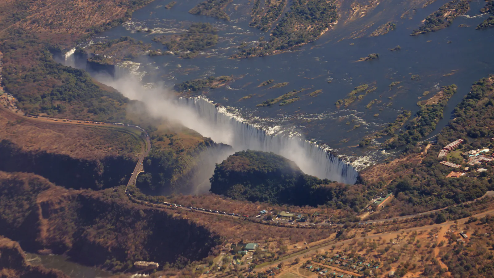 Aerial view of Victoria Falls: a long curtain of white water and mist along the lip of a deep gorge, with the Zambezi and its islands above and a road bridge crossing the gorge at left