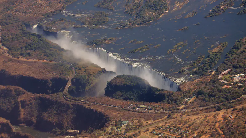 Aerial view of Victoria Falls: a long curtain of white water and mist along the lip of a deep gorge, with the Zambezi and its islands above and a road bridge crossing the gorge at left