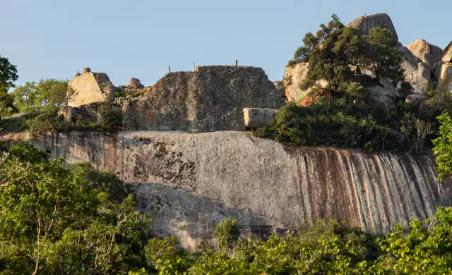 Dry-stone walls of the Hill Complex at Great Zimbabwe stand on top of a smooth granite cliff streaked with rust-coloured bands, among green trees and boulders under a clear sky
