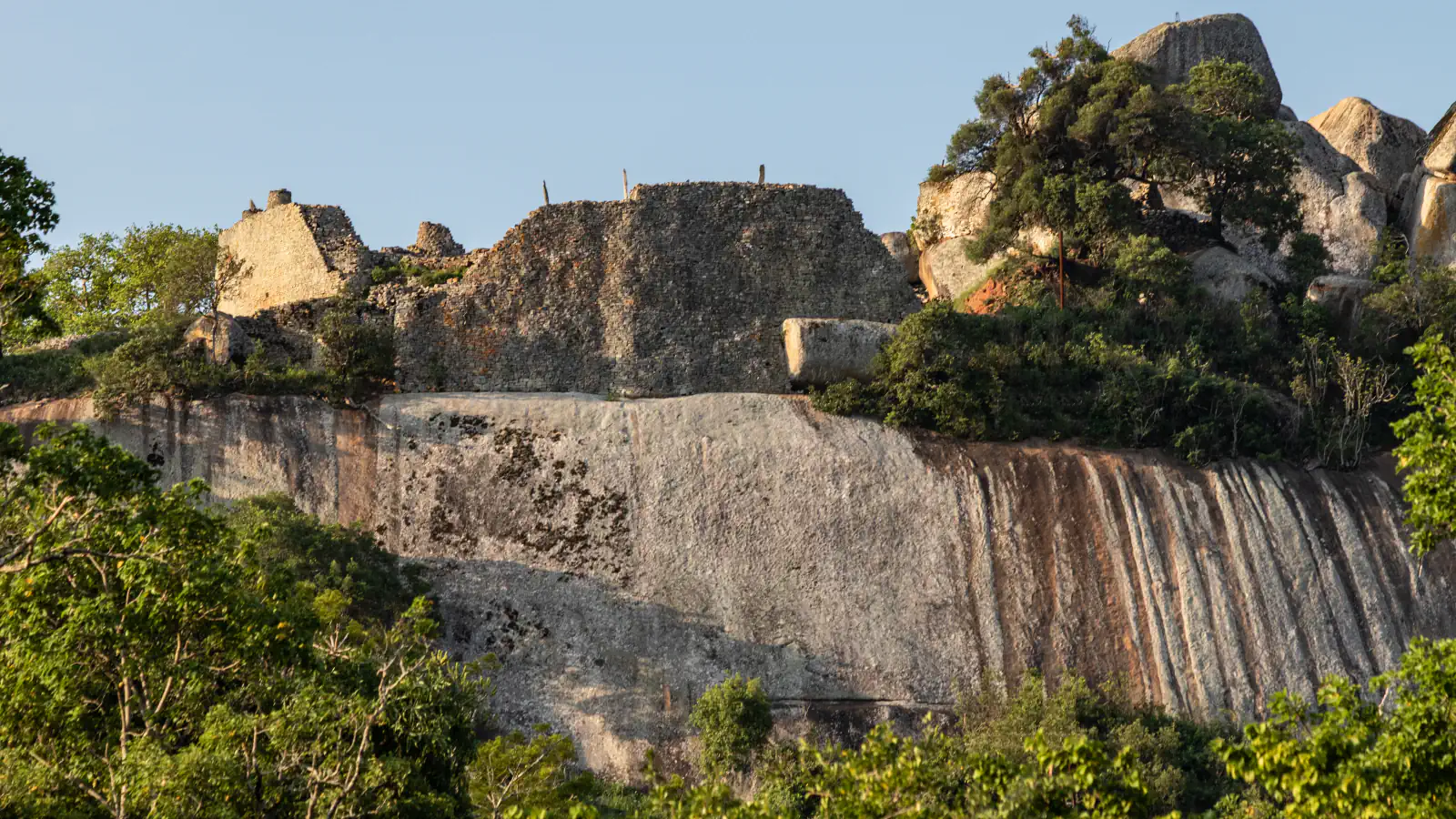 Dry-stone walls of the Hill Complex at Great Zimbabwe stand on top of a smooth granite cliff streaked with rust-coloured bands, among green trees and boulders under a clear sky
