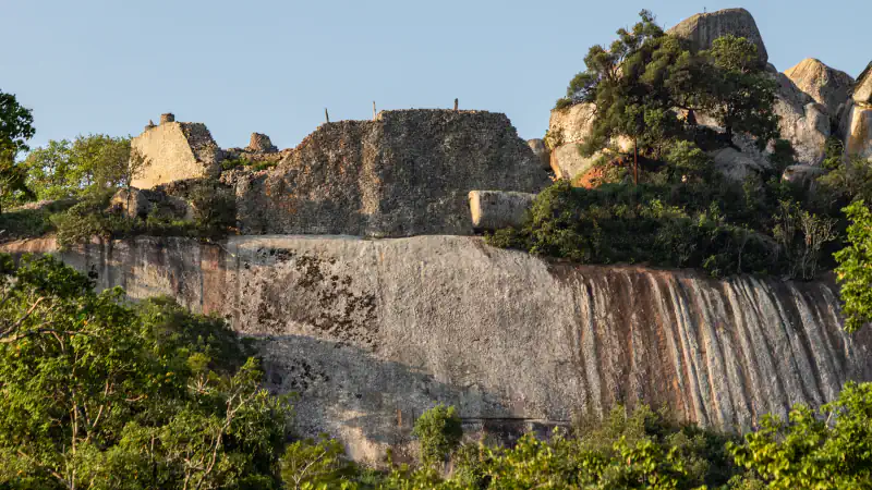 Dry-stone walls of the Hill Complex at Great Zimbabwe stand on top of a smooth granite cliff streaked with rust-coloured bands, among green trees and boulders under a clear sky